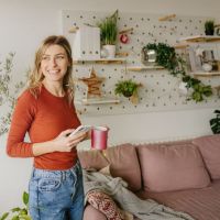 Frau steht mit einer Tasse Tee und Ihrem Handy in einem Wohnzimmer